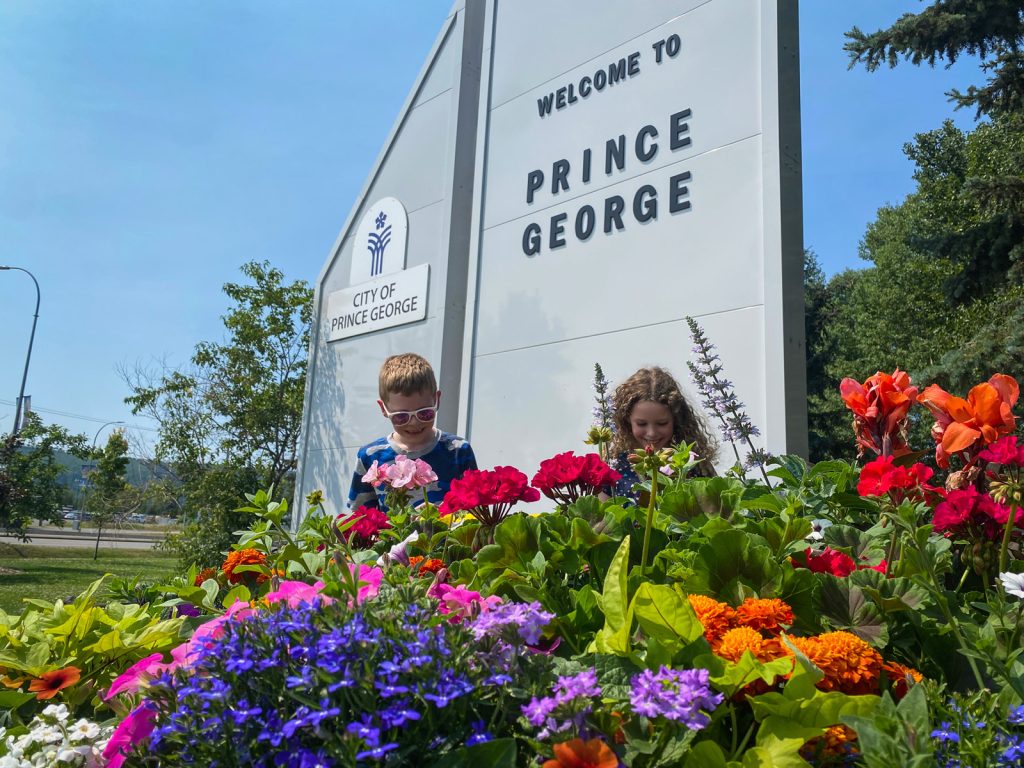 Two smiling children face the camera and look at a rainbow of colourful flowers in a large planter with the Welcome to Prince George sign in the background.